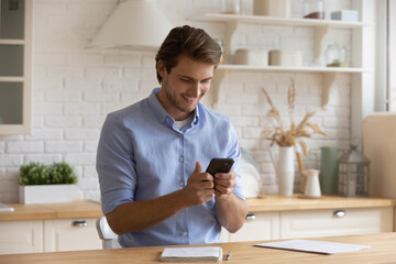 Smiling man holding smartphone, looking at screen, sitting at table in kitchen, having fun with mobile device apps, browsing surfing internet, typing writing message, shopping or chatting online