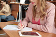© pressmaster - Girl with long blond hair making notes in copybook at lecture while sitting by desk against groupmates during individual work at lesson