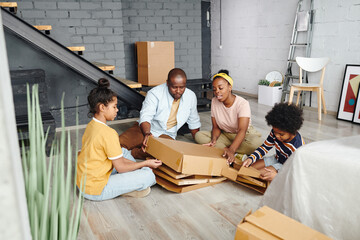 Canvas Print - Young African family sitting on the floor of large living-room in their new flat or house and unpacking cardboard box with furniture parts