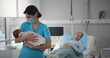 Nurse in medical mask holding newborn baby with mother resting in bed on background