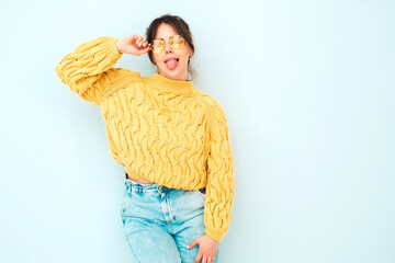 Wall Mural - Young beautiful smiling female in trendy summer yellow hipster sweater and  jeans. Sexy carefree woman posing near light blue wall in studio. Positive model having fun indoors. Shows tongue