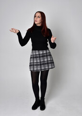 Wall Mural - Full length portrait of a red haired  girl wearing black turtleneck sweater,  plaid skirt and boots.  Standing pose against a studio background.