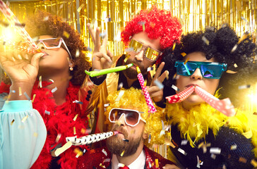 Multiethnic friends company in carnival wear blowing in party pipes have fun under confetti rain. Headshot portrait on nightclub sparkling background. Festive atmosphere, birthday, corporate event