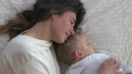 Poster - Authentic shot of young mother is caressing her toddler baby boy while sleeping by day on bed with soft blanket in a nursery. Concept of family, comfort,care, love,sweet dreams, motherhood, parenthood