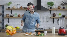 Handsome Joyful Middle Eastern Man In Headphone Cutting Tomato For Healthful Vegan Salad And Dancing. Portrait Of Cheerful Positive Millennial Having Fun Enjoying Music Cooking Healthy Food In Kitchen