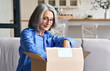 © insta_photos - Happy senior middle 60s aged woman opening box with ordered clothes at home on couch. Old mature online shopper customer opening online shop parcel. International delivery service comfort concept.