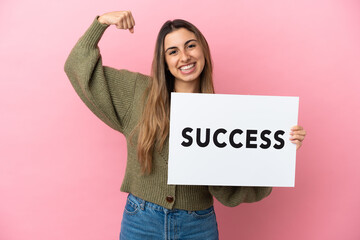 Wall Mural - Young caucasian woman isolated on pink background holding a placard with text SUCCESS and doing strong gesture