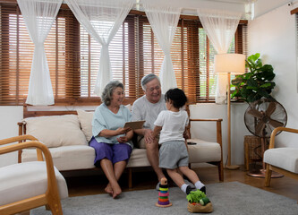 Happy Asian family spending time together at home. Grandfather and grandmother play with little grandchild girl in living room. Retirement senior couple having fun with cute baby granddaughter.