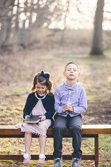 Wall Mural - Little Christian boy praying and a girl holding the holy bible while sitting on a bench in a park