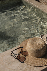 Sunglasses and straw hat on marble swimming pool side with clear blue water with waves sunlight shadow reflections. Minimal aesthetic summer vacation concept background.