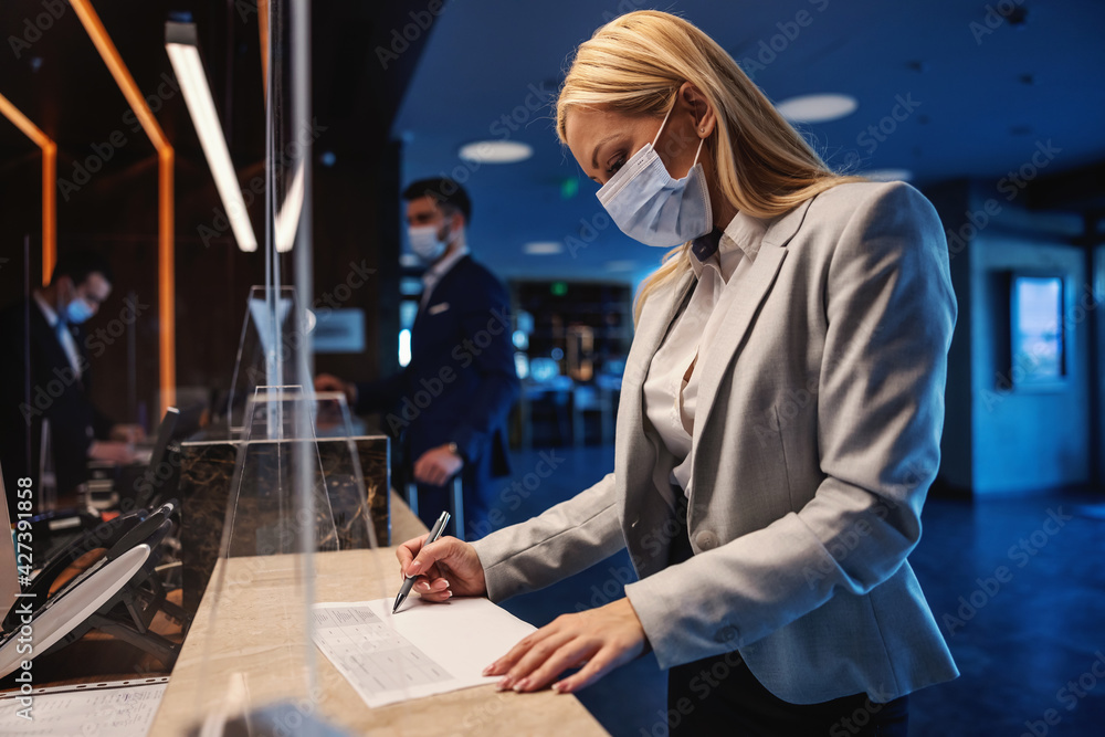 Blond businesswoman with face mask standing at reception in hotel and filling up the form during corona virus pandemic. Business trip, travel during corona, COVID19 precautionary measures - obrazy, fototapety, plakaty 