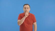 A Man In A Red T-shirt Looks From Side To Side, Then Somehow Asks For Silence And Points To A Stop. Indoor Studio Shot Isolated On Blue Background. 