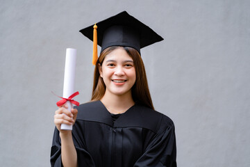 Wall Mural - A young happy Asian woman university graduate in graduation gown and mortarboard holds a degree certificate celebrates education achievement in the university campus.  Education stock photo