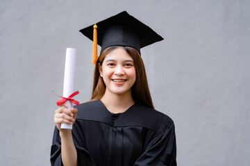 A young happy Asian woman university graduate in graduation gown and mortarboard holds a degree certificate celebrates education achievement in the university campus.  Education stock photo