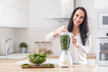 Beautiful lady blends greens in a smoothie mixer with more leaves inside the glass bowl next to her