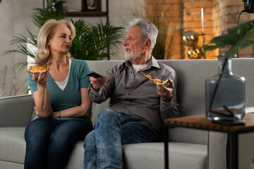 Wall Mural - Cheerful husband and wife sitting on sofa at home. Happy senior woman and man eating pizza while watching a movie..