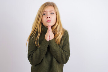 Wall Mural - Positive Caucasian kid girl wearing green knitted sweater against white wall smiles happily, glad to receive pleasant news from interlocutor, keeps palms together, People emotions concept.