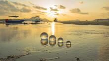 .The Golden Sun Rises Within The Glass Beads Arranged In A Shallow Canal, .the Water Gently Flows Through The Orb Into The Sea. .A Crystal Sphere Revealing The Upside-down Sunrise Sea View.