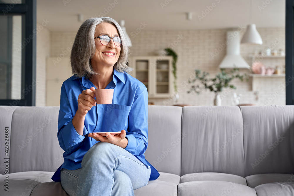 Smiling mature elder 60s woman sitting relaxing with cup of tea, coffee. Senior mid age stylish look woman with eyeglasses portrait with cup looking away at modern home. - obrazy, fototapety, plakaty 