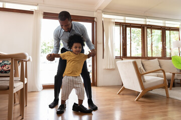 Wall Mural - Smiling African little child having fun stepping with his dad shoe in living room at home. Happy small African American kid playing with father. Loving Black Family. Curly hair in afro