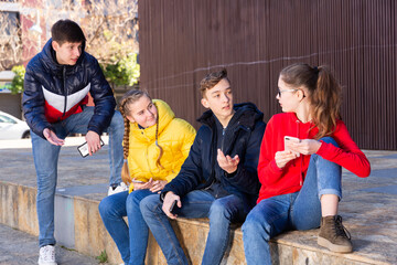  Four teenagers having friendly discussion and using cellphones during gathering outdoors on spring day