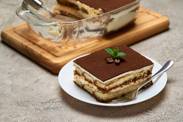 Traditional Italian Tiramisu dessert in glass baking dish and portion on grey concrete background