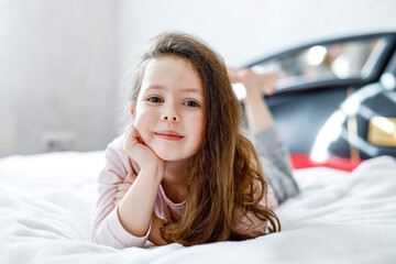 Adorable happy little kid girl with long hairs after sleeping in his white hotel bed in colorful nightwear. School child on family vacations. Funny happy child playing and smiling