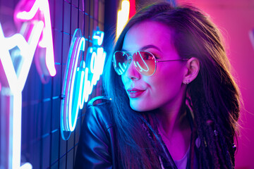 Fashion portrait of a young woman in sunglasses posing near neon signs in night club