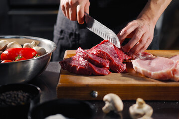 Wall Mural - The male chef cuts raw meat with a knife on a wooden board. The restaurant kitchen cooking.