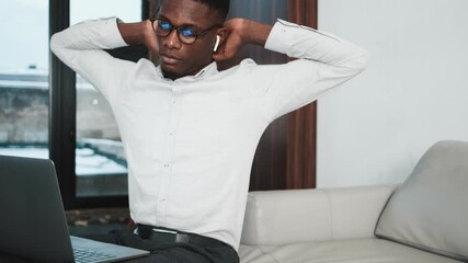 Poster - A concentrated african american businessman wearing earbuds is working with his laptop computer holding it on his knees while stretching his back sitting on the sofa inside the apartments