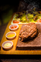 Poster - Grilled juicy beef steak smoking on hot stone served on wooden board with salad and sauces