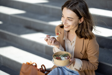 Pretty woman have outdoor lunch near office building while sitting on the stairs and smile. Healthy meal and coffee for takeaway. 