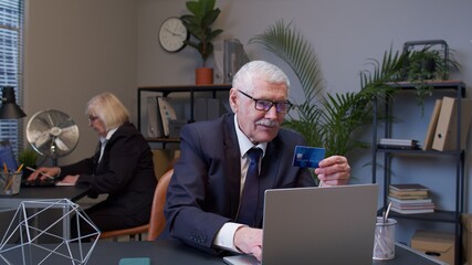 Senior businessman grandfather company director shopping online and paying with credit card in modern office. Elderly mature freelancer man buying on Internet enter credit card details. E-commerce