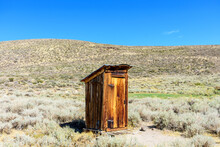 Old wooden outhouse in Bodie State Historic Park. Scenic rolling hills covered with sagebrush under blue sky.
