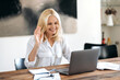 © Kateryna - Caucasian attractive elegant adult business woman in a stylish white shirt sitting on a workplace, uses laptop for communicate with colleagues by video conference, greeting interlocutor,friendly smile