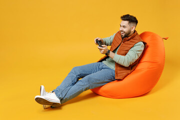 Wall Mural - Full length young gambling caucasian excited man 20s years old wear orange vest mint sweatshirt sitting in beanbag bag chair playing pc game with joystick console isolated on yellow background studio.