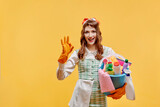 Fototapeta Tęcza - The happy cleaning lady shows the ok gesture and looks at the camera. A bottle of household chemicals and cleaning products. A sponge for washing dishes and a rag. Pin-up style. Yellow background.