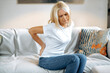 Upset frustrated mature woman suffering from backache, unhappy senior blonde sitting on a sofa at living room, feeling discomfort because of pain in back, have joint problems, need rest.