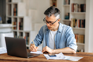 Focused mature gray haired intelligent caucasian businessman or freelancer with glasses, sitting at workplace, taking notes in notebook, studying or working online, using laptop