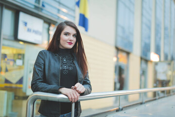 Poster - Young brunette female in a leather jacket posing leaning on a railing in a street