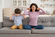 © DimaBerlin - Frowning mom and son sit on couch with closed eyes and cover ears from noisy music or fight sounds from neighbors. Young female parent and kid tired of drilling noise not listen plug ears with fingers