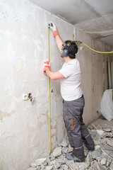 Wall Mural - Builder man using a measuring tape at the construction site. Construction worker wearing safety gloves, glasses and earmuffs.