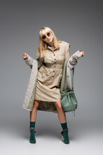Full Length Of Stylish Young Woman In Sunglasses And Trench Coat With Green Bag Posing On Grey