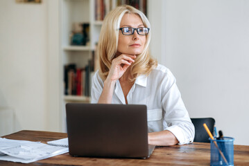 Attractive successful caucasian senior business woman, blond lady, wearing a white shirt and glasses, sits in front of laptop, taking break, looking pensively to the side, dreaming about vacation