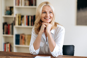 Portrait of happy elegant successful mature blonde woman in white stylish shirt, sits at the desk at office, taking a break from work, looking to the side, thinking, smiling friendly