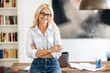 © Kateryna - Portrait of happy successful elegant middle aged caucasian woman, stylishly dressed, with glasses, standing near the table, with arms crossed, looking at camera, smiling friendly.