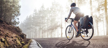 Cyclist On A Bicycle With Panniers Riding Along A Foggy Forest Road