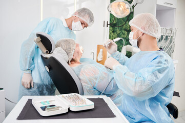 Wall Mural - Horizontal snapshot in dentist office of dentist and his assistant during surgery performance. People wearing disposable closing , gloves, caps and masks. Surgical taper kit on the table.