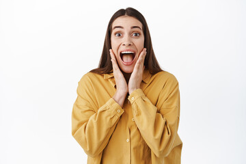 Image of impressed young woman looking at something fascinated and unbelievable, scream with joy in awe, touching cheeks and drop jaw astonished, white background