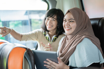 Wall Mural - two young women smiling with pointing fingers while chatting while holding a tablet sitting on the bus